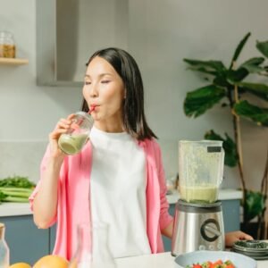 Asian woman sipping a green smoothie in a modern kitchen setting.